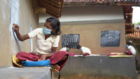 A student writing on a blackboard in the wall outside a mud house